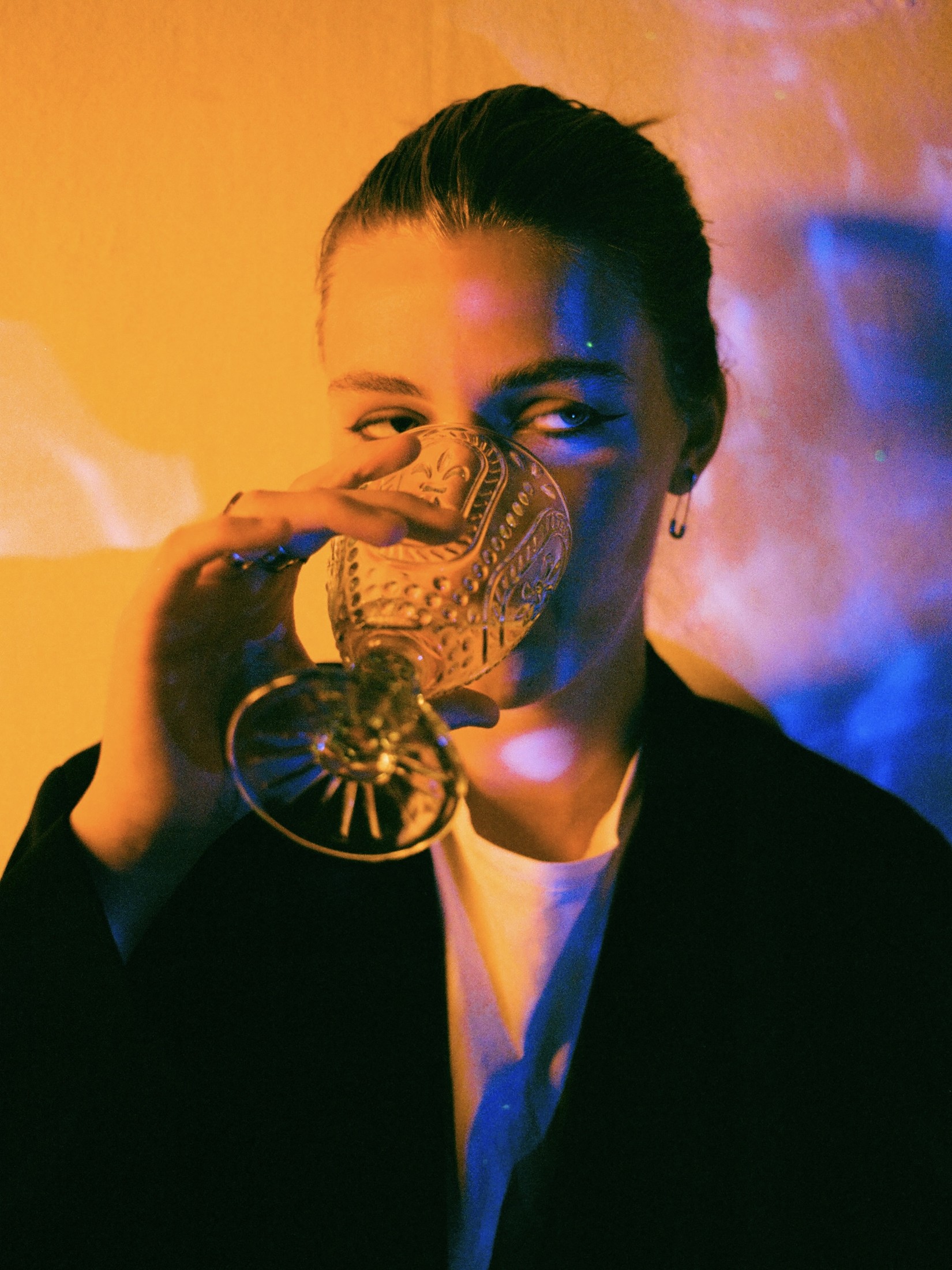 Close-up portrait lit in blue and amber tones, sipping from an ornate crystal glass
